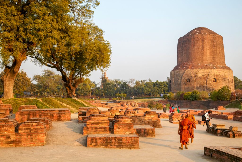 Sarnath: Buddhist monks walk among ancient brick ruins and Dhamek Stupa under large trees in historic pilgrimage site near Varanasi, India.