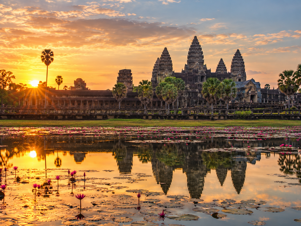 Angkor Wat temple in Siem Reap, Cambodia, glowing at sunrise with palm trees, lotus-filled water, and a clear reflection of the historic temple complex.