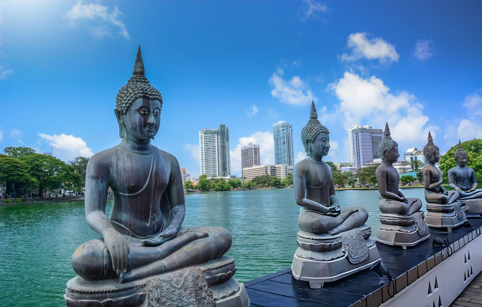 Seated Buddha statues beside Beira Lake in Colombo, Sri Lanka, with modern city buildings, green trees, and a bright blue sky in the background.