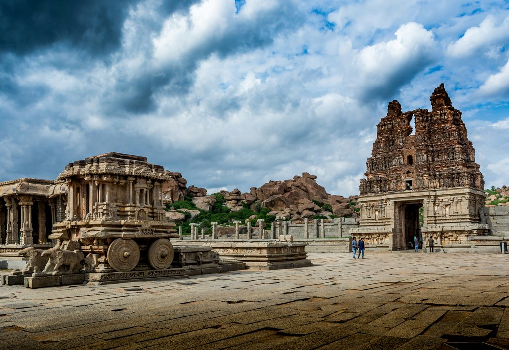 The stone chariot and grand gopuram (tower) of the Vittala Temple complex in Hampi, Karnataka, under a dramatic cloudy sky, highlighting Underrated Places in India.