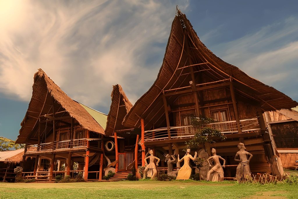 Traditional Mishing tribal stilt houses with large thatched roofs on Majuli Island, Assam, featuring cultural statues in front, one of the Underrated Places in India.