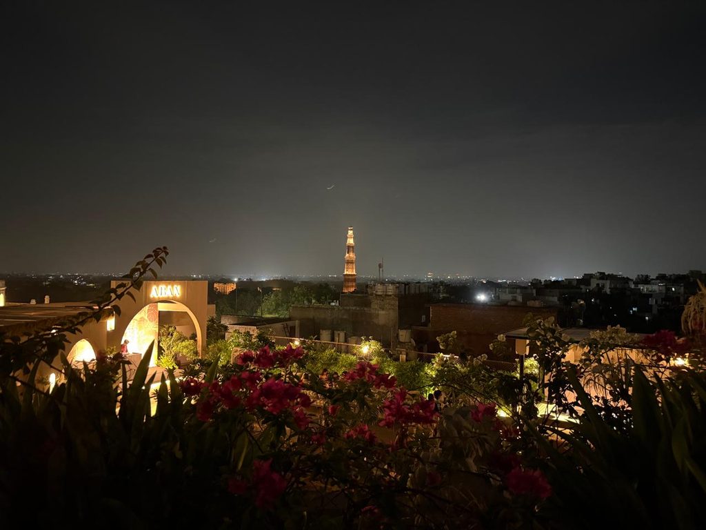 Night view from a rooftop bar in Delhi with illuminated Qutub Minar in the distance, city lights, garden greenery, and a dark evening skyline.