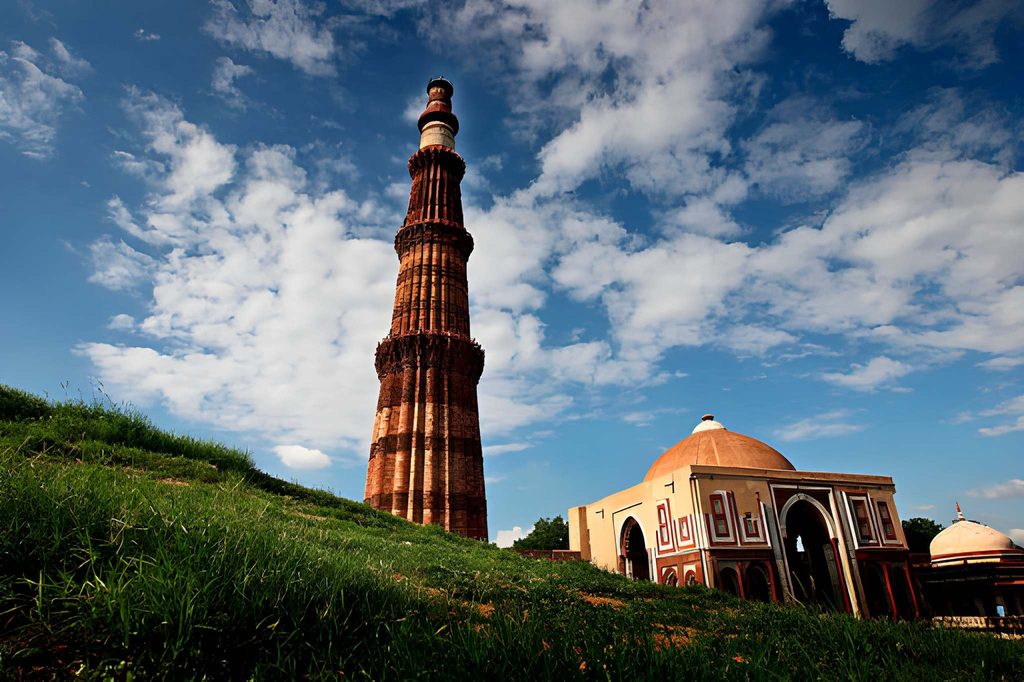 Qutub Minar in Delhi rising beside historic Indo-Islamic architecture, surrounded by green lawns under a bright blue sky with scattered clouds.
