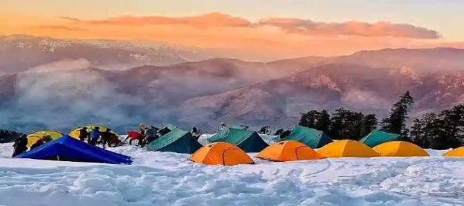 Snow-covered Kedarkantha trek campsite with colorful tents overlooking Himalayan mountains at sunrise.
